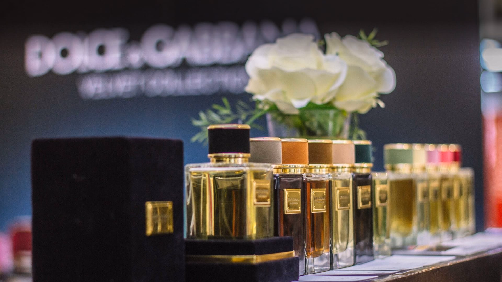 A row of gold-capped fragrance bottles receding into soft focus beside white roses in a ribbed green vase, on a dark counter