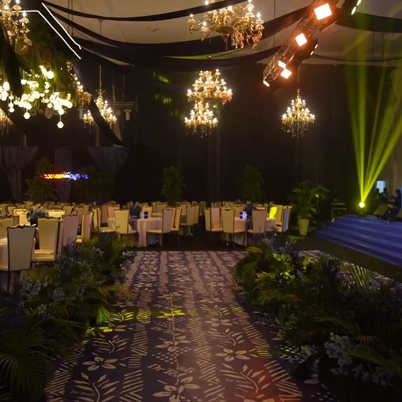A dark banquet hall lit by chandeliers, with a patterned runway leading to a green-lit stage