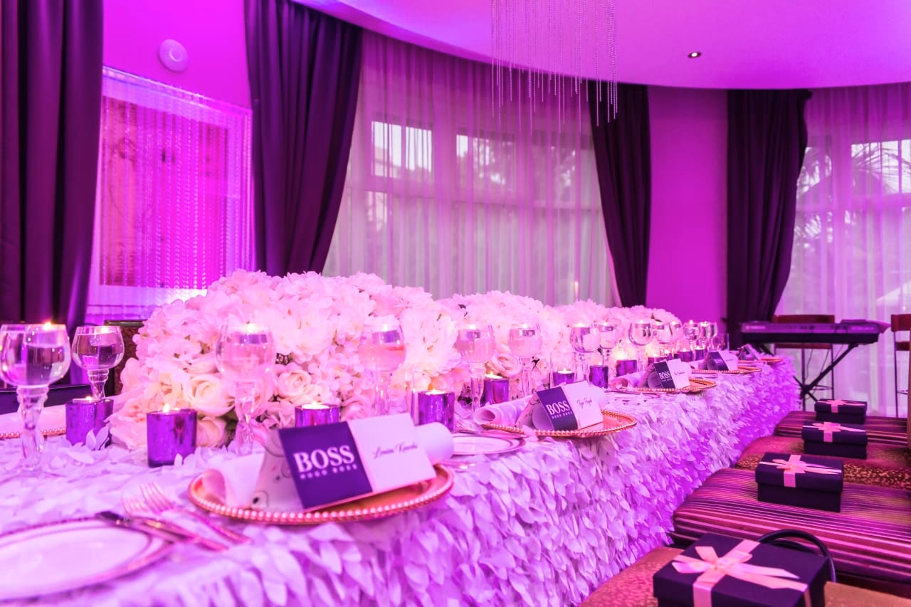 A long banquet table under magenta uplighting, dressed with a dense runner of pale flowers, BOSS place cards and gold-rimmed chargers, with gift boxes at each setting