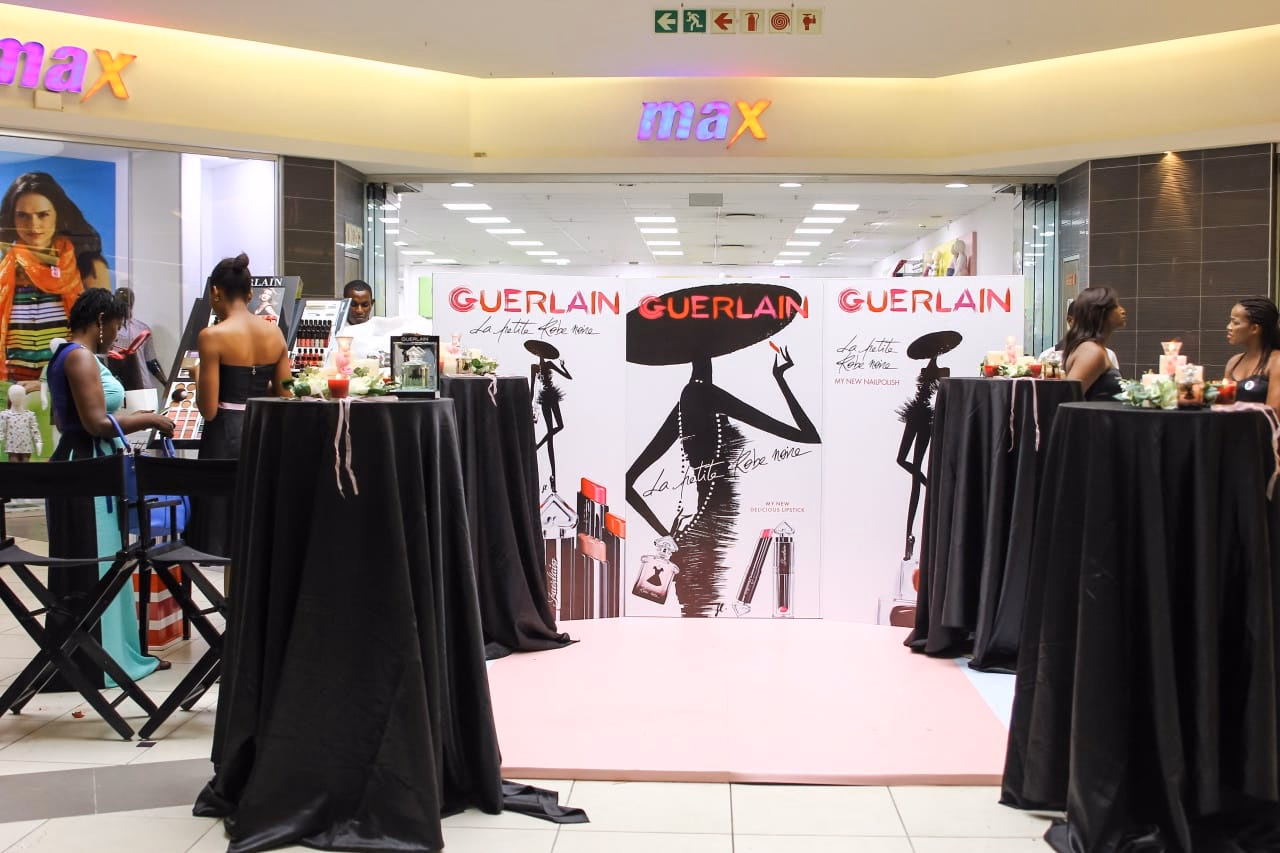 A Guerlain event space with black-draped cocktail tables on a pink carpet, guests standing at them, and a branded backdrop carrying the Guerlain silhouette