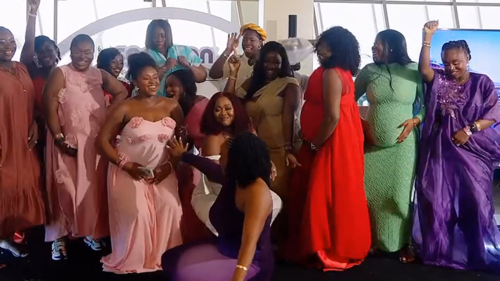 A large group of women in brightly coloured dresses standing together in a bright room with tall windows and a blue patterned carpet, facing the camera