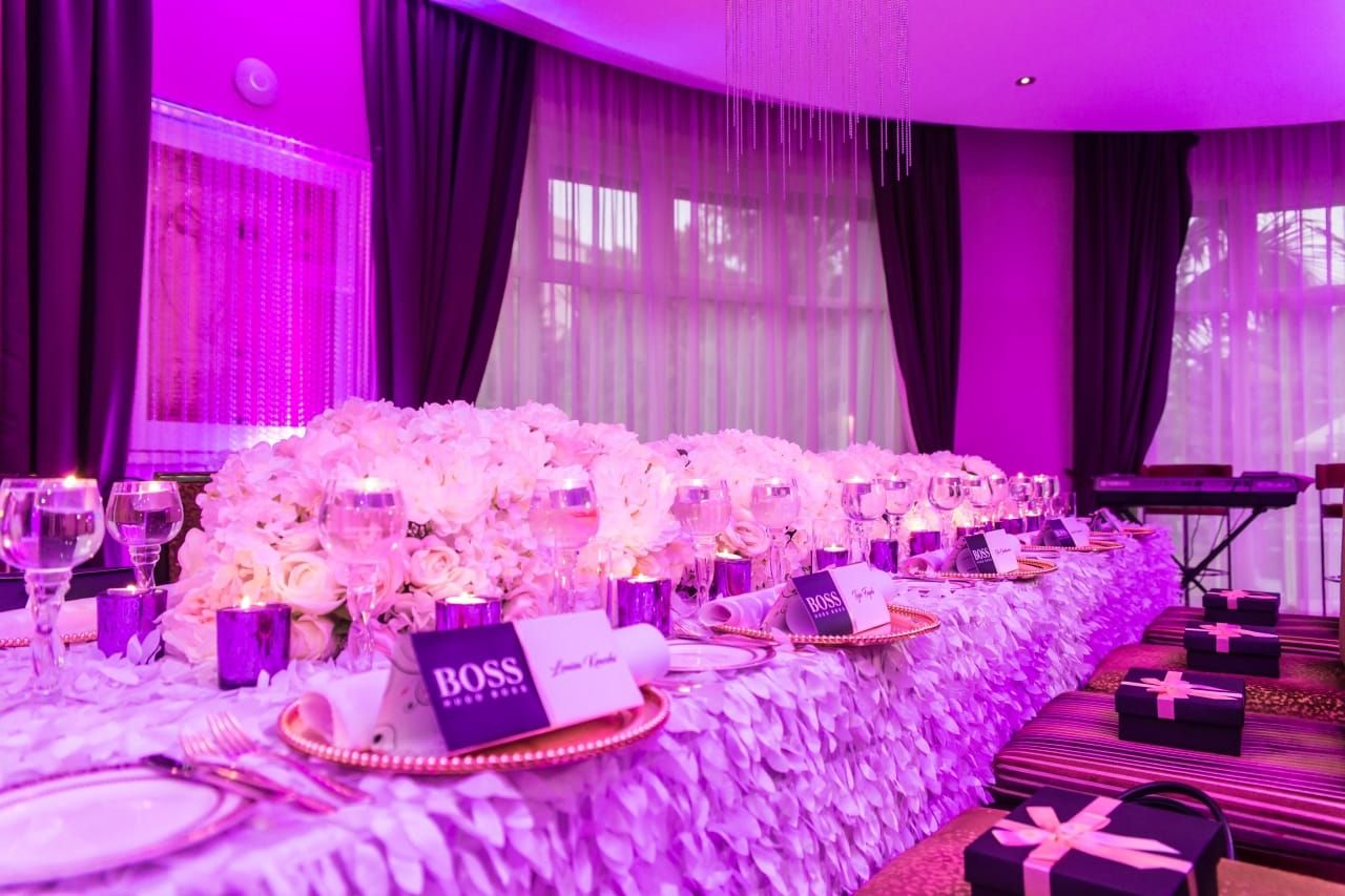 A long banquet table under magenta uplighting, dressed with a dense runner of pale flowers, BOSS place cards and gold-rimmed chargers, with gift boxes at each setting