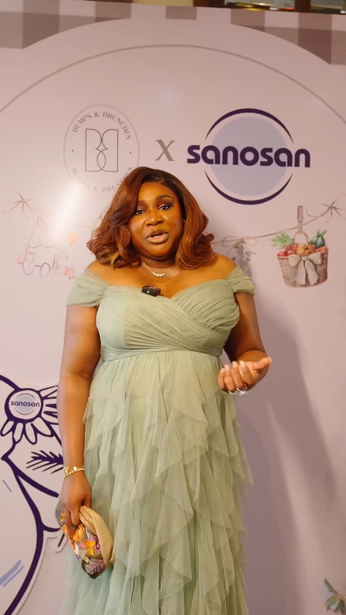 A guest in a pale green dress standing in front of a printed backdrop carrying the Sanosan wordmark beside a partner's circular mark, over photography of tulle dresses and a line drawing of a rabbit